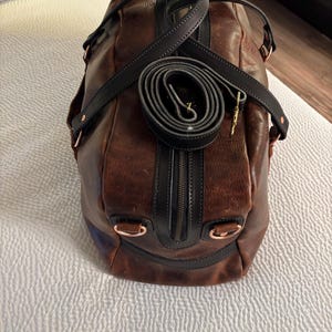 Op de afbeelding: Een bruine leren reistas met zwarte leren afwerking, een centrale ritssluiting en koperkleurige metalen hardware. Een opgerolde zwarte schouderriem met een goudkleurige metalen clip ligt bovenop de tas, die is voorzien van D-ringen aan de zijkant.