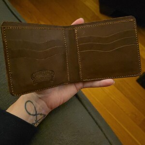 May include: A brown leather bifold wallet featuring multiple card slots with wavy edges and visible stitching. The interior is embossed with the text Corbett Leather Works. The wallet is open to display the internal compartments and pockets.