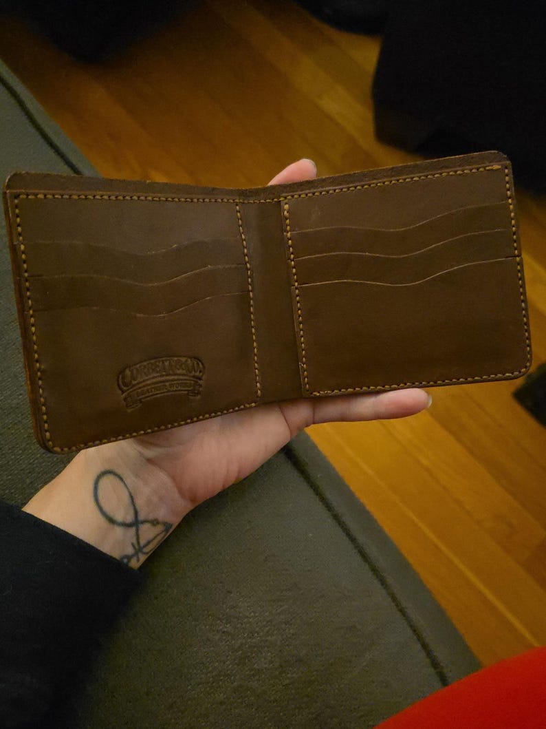 May include: A brown leather bifold wallet featuring multiple card slots with wavy edges and visible stitching. The interior is embossed with the text Corbett Leather Works. The wallet is open to display the internal compartments and pockets.