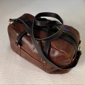 Op de afbeelding: Een donkerbruine leren reistas met zwarte leren handvatten en een zwarte leren schouderriem. De tas is voorzien van een ritssluiting aan de bovenkant, koperkleurige metalen klinknagels en een rechthoekige structuur met zichtbare stiksels.