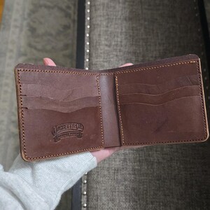 May include: A brown leather bifold wallet featuring multiple card slots and a debossed logo that reads "CORBEAT & CO. LEATHER WORKS". The wallet is constructed with visible stitching along the edges and has a smooth, matte finish.