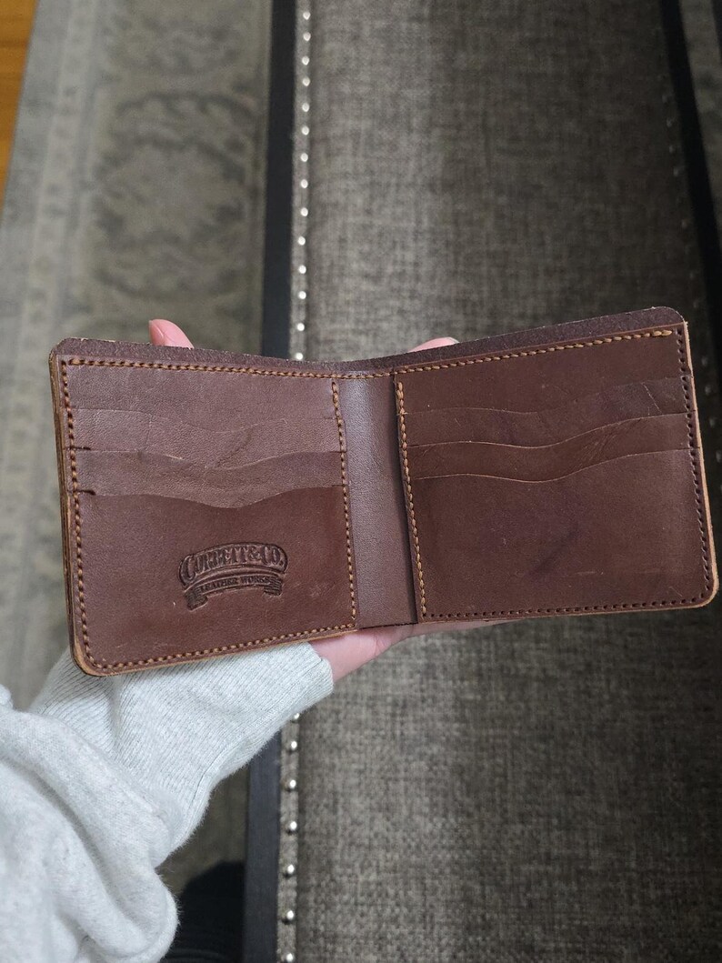 May include: A brown leather bifold wallet featuring multiple card slots and a debossed logo that reads "CORBEAT & CO. LEATHER WORKS". The wallet is constructed with visible stitching along the edges and has a smooth, matte finish.