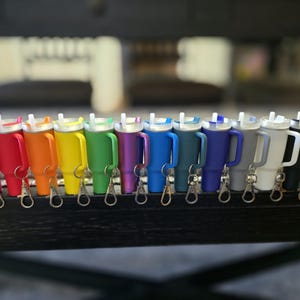 May include: A collection of colorful insulated tumblers with handles and attached keychains. The tumblers are arranged in a row, showcasing a rainbow of colors including pink, red, orange, yellow, green, purple, blue, and black.