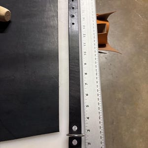 May include: A black leather strap with a silver metal buckle and a D-ring, featuring six punched holes at one end. The strap is positioned next to a metal ruler showing a length of approximately 20 inches from the buckle to the end.