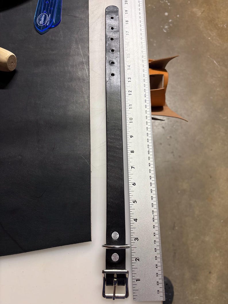May include: A black leather strap with a silver metal buckle and a D-ring, featuring six punched holes at one end. The strap is positioned next to a metal ruler showing a length of approximately 20 inches from the buckle to the end.