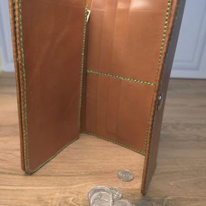 Puede incluir: Una cartera larga de cuero color canela con costuras de color verde brillante a lo largo de los bordes y bolsillos interiores. La cartera est&aacute; abierta, revelando varias ranuras para tarjetas y un compartimento con cremallera.