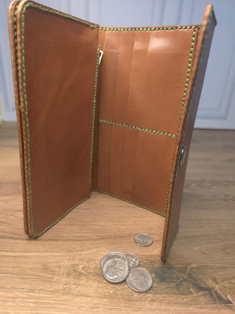Puede incluir: Una cartera larga de cuero color canela con costuras de color verde brillante a lo largo de los bordes y bolsillos interiores. La cartera est&aacute; abierta, revelando varias ranuras para tarjetas y un compartimento con cremallera.