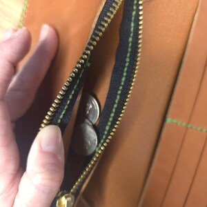 Puede incluir: Una cartera de cuero color canela con un bolsillo monedero negro con cremallera y tirador de metal dorado. La cartera est&aacute; confeccionada con costuras de color verde brillante en los bordes e incluye varias ranuras para tarjetas.