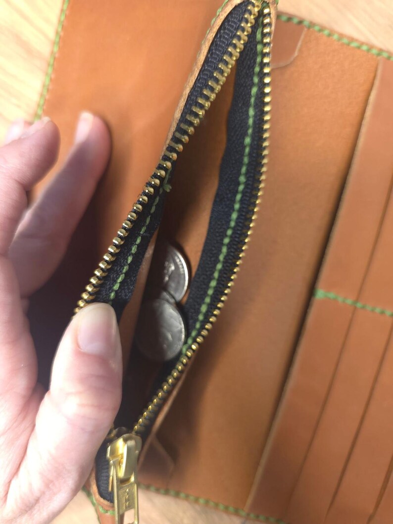 Puede incluir: Una cartera de cuero color canela con un bolsillo monedero negro con cremallera y tirador de metal dorado. La cartera est&aacute; confeccionada con costuras de color verde brillante en los bordes e incluye varias ranuras para tarjetas.