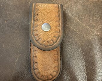 May include: A tan leather pouch with a basketweave texture and a scalloped edge border. The pouch features a centered silver-toned metal snap closure and is constructed with dark brown stitching along the perimeter of the flap and body.