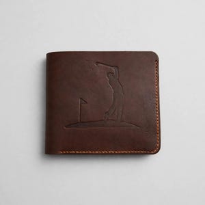 May include: A dark brown leather bifold wallet featuring an embossed graphic of a golfer swinging a club on a green with a flag. The wallet is constructed with visible orange-brown stitching along the edges and a smooth matte finish.