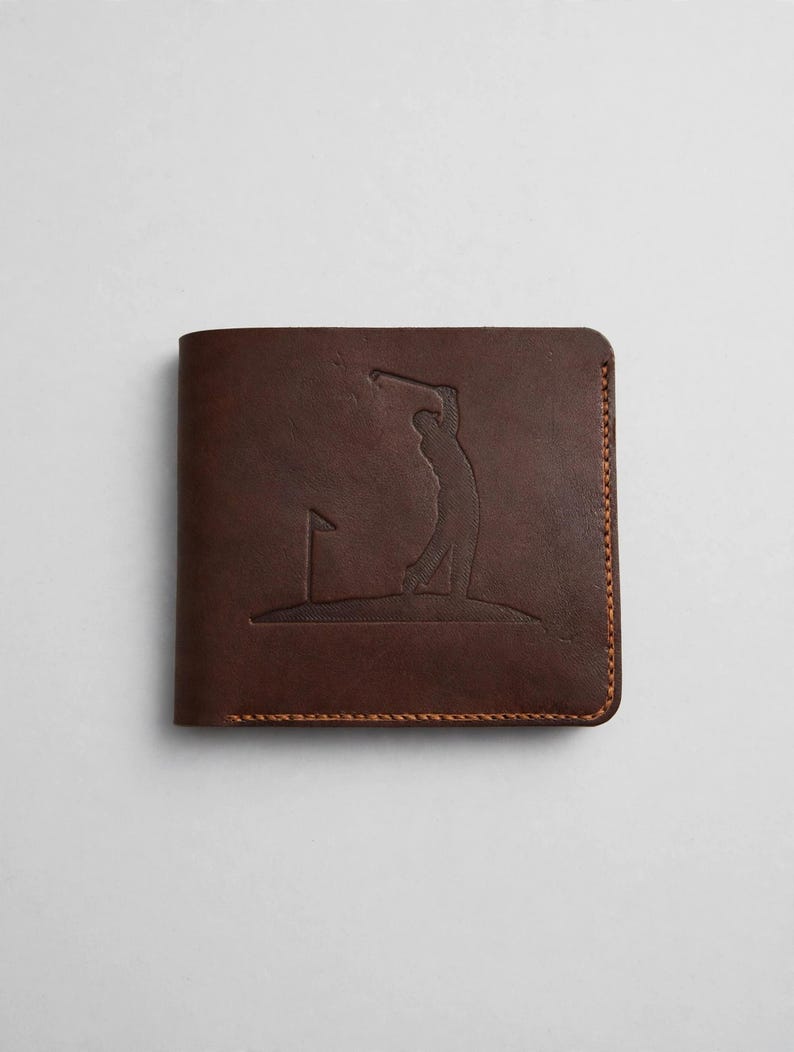May include: A dark brown leather bifold wallet featuring an embossed graphic of a golfer swinging a club on a green with a flag. The wallet is constructed with visible orange-brown stitching along the edges and a smooth matte finish.