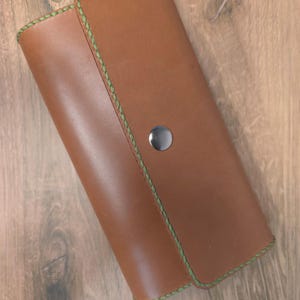 Puede incluir: Una cartera de cuero color canela con cierre de solapa rectangular y esquinas redondeadas. La cartera presenta costuras verdes visibles a lo largo de los bordes y se asegura con un bot&oacute;n de presi&oacute;n central de metal plateado.