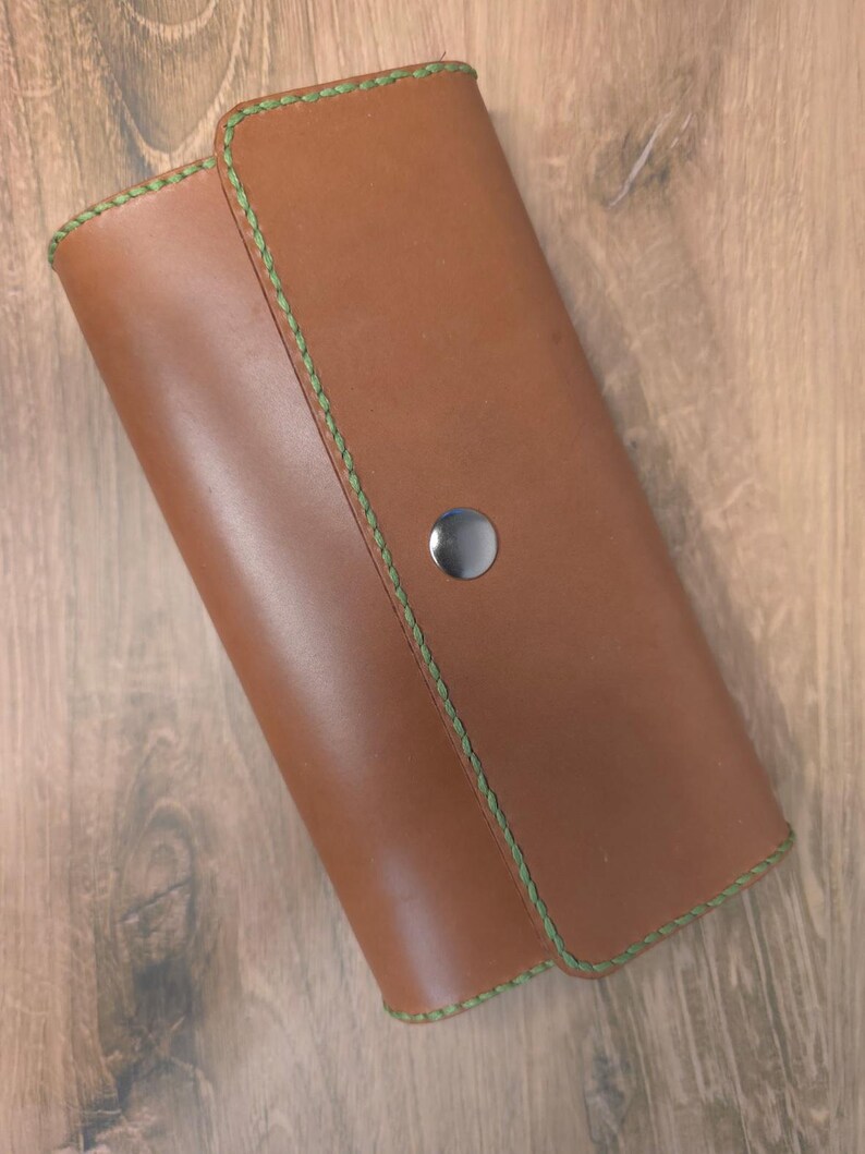 Puede incluir: Una cartera de cuero color canela con cierre de solapa rectangular y esquinas redondeadas. La cartera presenta costuras verdes visibles a lo largo de los bordes y se asegura con un bot&oacute;n de presi&oacute;n central de metal plateado.