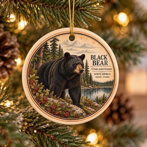 May include: A round ceramic ornament featuring a detailed illustration of a black bear standing on a rocky, berry-covered slope near a lake and forest. The text reads BLACK BEAR, Ursus americanus, NORTH AMERICA, and Family: Ursidae in black font.