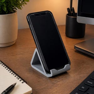 Könnte beinhalten: Ein grauer, dreieckiger Telefonständer, der ein schwarzes Smartphone hält. Der Ständer steht auf einem Holzschreibtisch, auf dem ein Notizbuch, ein Stift und eine Tastatur zu sehen sind. Eine kleine Pflanze und eine Schreibtischlampe befinden sich im Hintergrund.