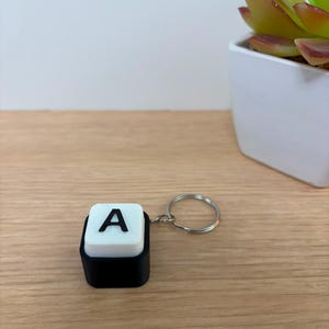 May include: A keychain featuring a square, black base topped with a white, raised square button. The button displays a bold, black capital letter A in the center. A silver-toned metal split ring is attached to the side of the black base.