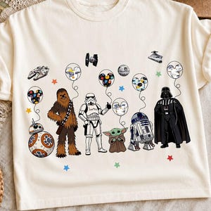 May include: A cream-colored t-shirt featuring a graphic print of Star Wars characters including Chewbacca, a Stormtrooper, Grogu, R2-D2, Darth Vader, and BB-8. The characters are holding balloons shaped like Mickey Mouse ears, interspersed with small colorful stars.