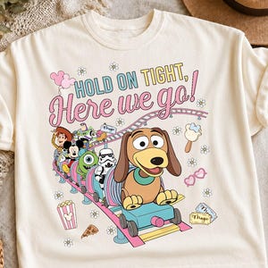 May include: A cream-colored t-shirt featuring a colorful graphic of various cartoon characters riding a roller coaster. The text reads, "HOLD ON TIGHT, Here we go!" The design includes small flowers, a popcorn bucket, a slice of pizza, and tickets.