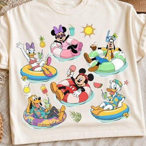 May include: A cream-colored t-shirt featuring a graphic print of Disney characters including Mickey Mouse, Minnie Mouse, Donald Duck, Daisy Duck, Goofy, and Pluto floating in colorful inner tubes, holding drinks and ice cream, with a sun icon.