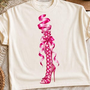 May include: A cream-colored t-shirt featuring a graphic print of a bright pink high-heeled sandal. The sandal has a tall, lace-up design that extends up the leg, adorned with flowing pink ribbons and a large bow tied at the center of the laces.