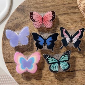 May include: A collection of six butterfly-shaped phone grips in various colors and designs. The grips feature pink, blue, and rainbow patterns. The butterflies are set against a wooden background.