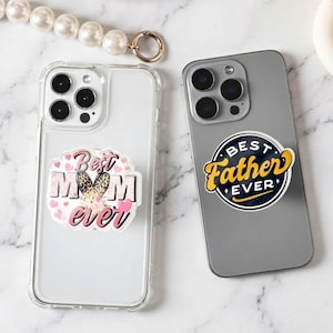 May include: Two smartphones, one in a clear case with a "Best Mom Ever" sticker and the other with a "Best Father Ever" sticker. The stickers have a pink and black color scheme. A pearl necklace and gold ring are in the background.