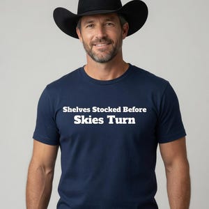 May include: A navy blue short-sleeved t-shirt featuring the white text "Shelves Stocked Before Skies Turn" printed across the chest. The shirt is paired with a black cowboy hat, which has a wide brim and a high, pinched crown design.