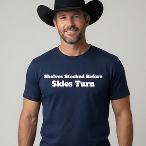 May include: A navy blue short-sleeved t-shirt featuring the white text "Shelves Stocked Before Skies Turn" printed across the chest. The shirt is paired with a black cowboy hat, which has a wide brim and a high, pinched crown design.