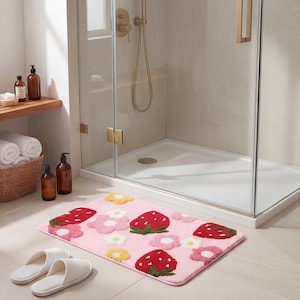 Puede incluir: Una alfombra de baño rosa con un estampado de fresas y flores. La alfombra presenta fresas rojas grandes con copas verdes y flores blancas, rosas y amarillas. Hay zapatillas blancas en primer plano. La alfombra está frente a una ducha de cristal.