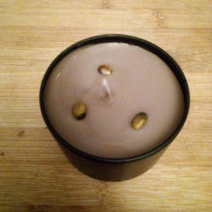 May include: A round, lavender-colored candle in a black metal container. The candle has three decorative gold-colored accents and a single wick. The candle sits on a wooden surface.