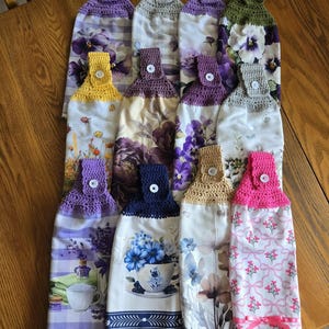 May include: A collection of twelve hanging kitchen towels, each featuring a crocheted top in various solid colors including purple, yellow, blue, pink, and green, secured with a white button. The towels display diverse floral and botanical prints.