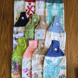 May include: A collection of twelve hanging kitchen towels, each featuring a crocheted top in various solid colors with a white button closure, attached to a fabric towel with diverse printed patterns including floral, animal, and seasonal designs.