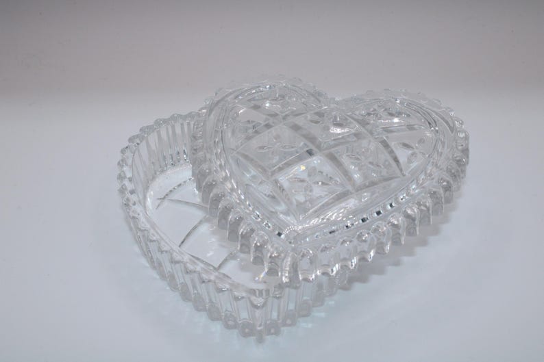 Pu&ograve; includere: Un portagioie in vetro trasparente a forma di cuore con coperchio rimovibile. Il coperchio presenta un motivo a griglia pressata con dettagli floreali, mentre la base e i bordi sono decorati con un design a coste verticali e un bordo perlato.