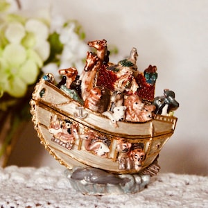 Può includere: Una statuetta decorativa dell'arca di Noè con una barca color crema e finiture dorate, riempita con varie figure di animali dipinte a mano tra cui giraffe, leoni e uccelli, con una struttura del tetto rossa e un dettaglio ancora sul lato.