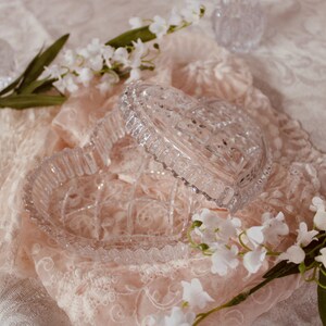 Pu&ograve; includere: Un portagioie trasparente a forma di cuore con coperchio, appoggiato su un tessuto di pizzo rosa. Fiori bianchi e verde circondano la scatola, creando un'estetica morbida e romantica. Lo sfondo &egrave; un tessuto bianco fantasia.