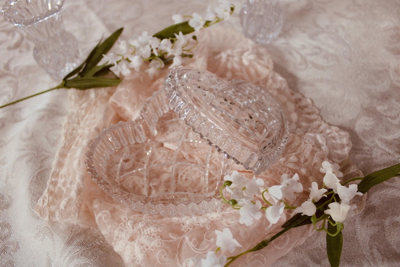 Pu&ograve; includere: Un portagioie trasparente a forma di cuore con coperchio, appoggiato su un tessuto di pizzo rosa. Fiori bianchi e verde circondano la scatola, creando un'estetica morbida e romantica. Lo sfondo &egrave; un tessuto bianco fantasia.
