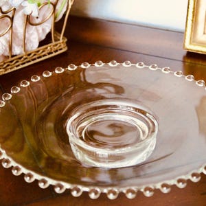 May include: A clear glass serving bowl featuring a wide, flat rim decorated with a continuous row of small, raised glass beads along the outer edge. The center of the bowl is recessed, creating a circular well with smooth, transparent glass walls.