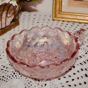 Può includere: Una piccola ciotola rotonda in vetro rosa iridescente con un motivo a rombi testurizzato sull'esterno e un bordo smerlato. La ciotola include una singola piccola maniglia su un lato, con una finitura riflettente che cattura la luce per mostrare sottili tonalità arcobaleno.