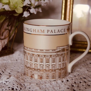 May include: A white ceramic mug featuring a gold-toned illustration of Buckingham Palace on the lower half. The upper section has a soft cream band with the text BUCKINGHAM PALACE printed in gold lettering, accompanied by a small gold crown icon.