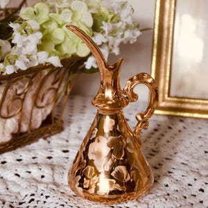 May include: A metallic gold ceramic pitcher with a conical body and a curved handle. The surface features a repeating pattern of light-colored floral shapes. The pitcher has a narrow, elongated spout extending upward from the top opening.