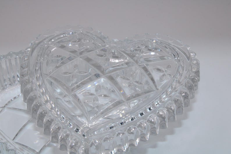 Pu&ograve; includere: Un piatto in vetro trasparente a forma di cuore con un motivo geometrico sfaccettato sul coperchio. I bordi del piatto sono decorati con una serie di creste verticali arrotondate, creando un bordo strutturato attorno al cuore.