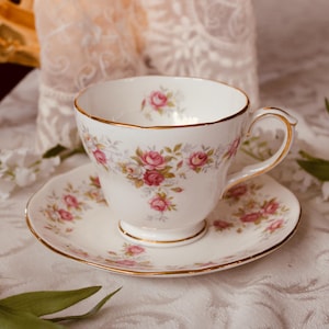 Pode incluir: Uma chávena e pires brancos com um padrão floral de rosas e detalhes dourados. A chávena está sobre o pires a condizer. O conjunto é rodeado por flores brancas e vegetação, criando uma estética delicada e elegante.