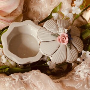 Può includere: Un piccolo portagioie in ceramica color bianco sporco con bordo smerlato e coperchio abbinato. Il coperchio ha la forma di un fiore con petali a coste ed è sormontato da una piccola rosa rosa scolpita, un minuscolo fiore bianco e una foglia blu.