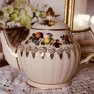 Peut inclure: Une théière en céramique couleur crème avec une base cannelée et des finitions dorées sur l'anse, le bec et le couvercle. Le corps est orné d'un motif de fruits colorés, poires et baies, avec un décor en filigrane doré sur le milieu.