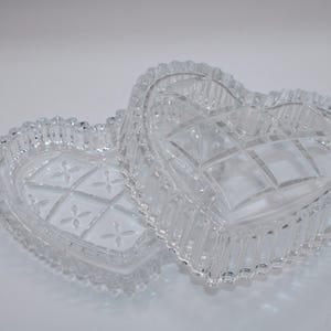Pu&ograve; includere: Un portagioie in vetro trasparente a forma di cuore con coperchio rimovibile. La base e il coperchio presentano un motivo geometrico pressato con piccoli disegni floreali al centro, rifinito con un bordo smerlato e perlato.