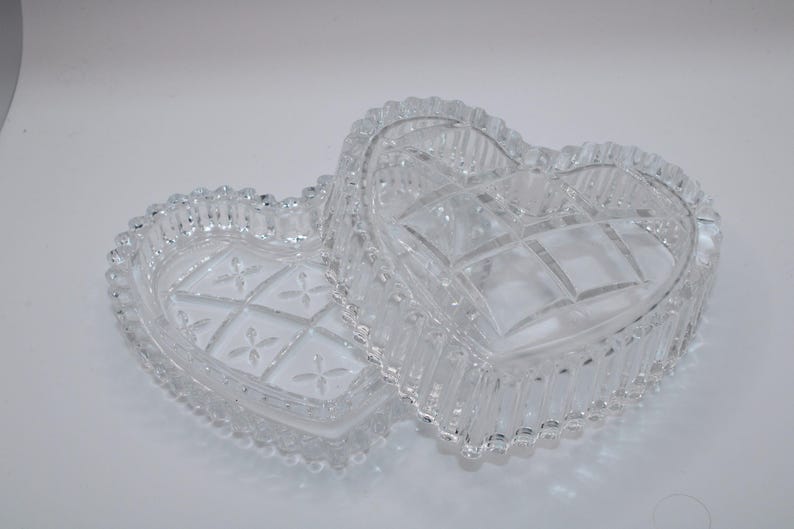 Pu&ograve; includere: Un portagioie in vetro trasparente a forma di cuore con coperchio rimovibile. La base e il coperchio presentano un motivo geometrico pressato con piccoli disegni floreali al centro, rifinito con un bordo smerlato e perlato.