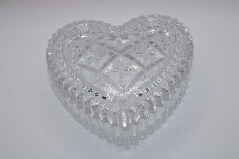 Pu&ograve; includere: Un portagioie trasparente a forma di cuore. La scatola &egrave; realizzata in cristallo tagliato con un motivo geometrico dettagliato sul coperchio e sui lati. I bordi hanno un design smerlato. La scatola &egrave; vuota e poggia su una superficie bianca.
