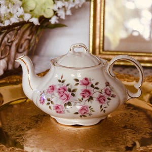 Puede incluir: Una tetera de porcelana color crema con un delicado patrón floral de rosas rosadas, hojas verdes y bordes dorados en el pico, el asa y la tapa. La tetera tiene un diseño de borde festoneado y se asienta sobre una bandeja de metal dorada.