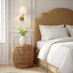 May include: A bedroom scene featuring a round, woven rattan side table with a white vase holding white calla lilies. A bed with white bedding and a gold-toned headboard is in the background. A gold wall sconce is mounted on the wall.